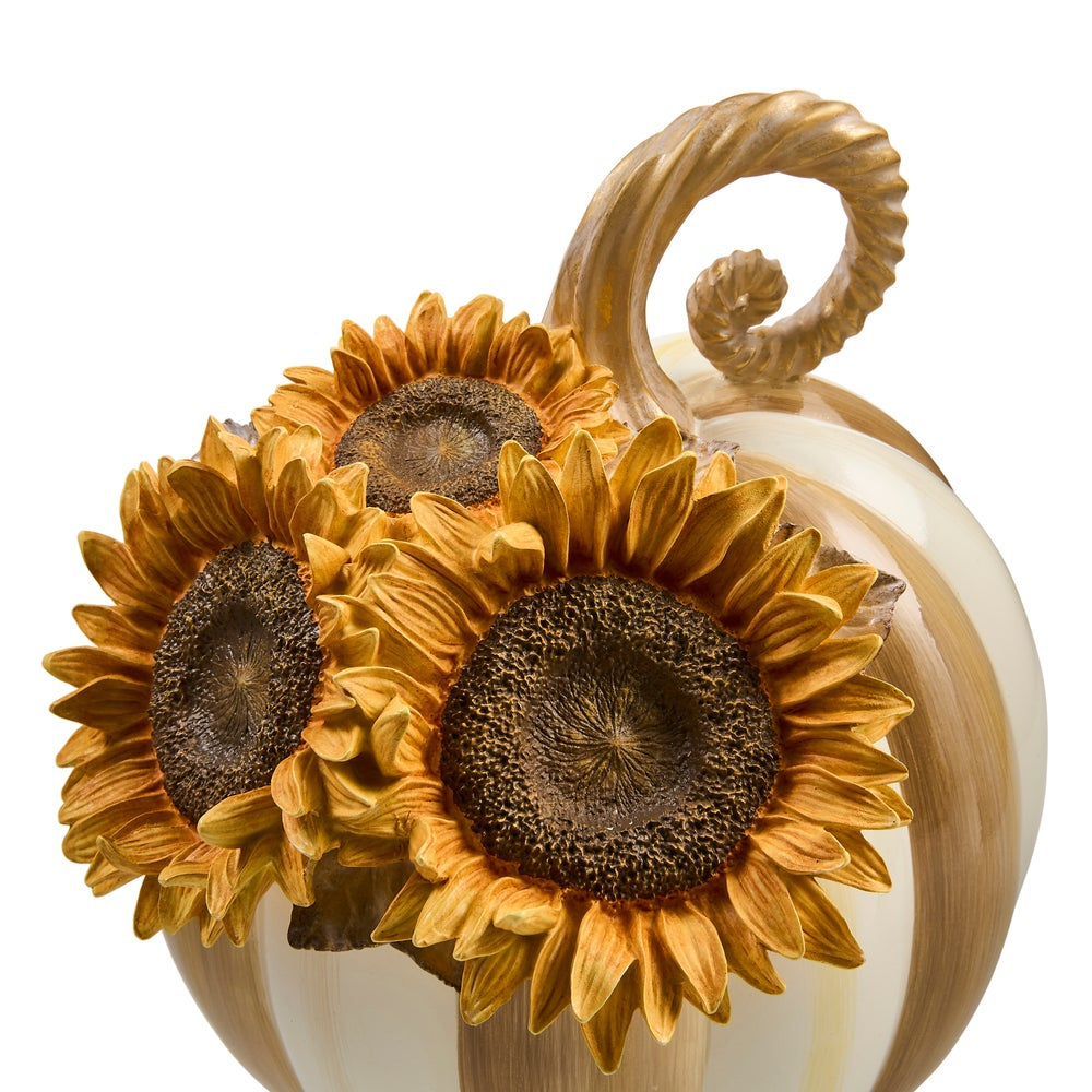 A decorative pumpkin sculpture with a twisted handle, painted in warm cream and tan stripes, adorned with three large golden sunflower decorations on the front, set against a plain white background.