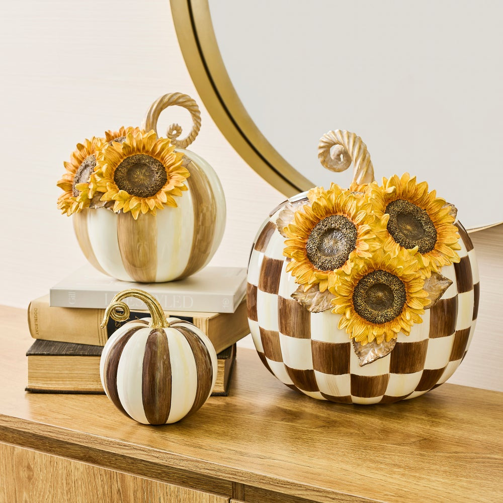 Three decorative pumpkins arranged on a wooden surface with stacked books and a round mirror in the background. The pumpkins have autumnal patterns: one is cream and tan with yellow sunflowers, one is brown and cream checkered with yellow sunflowers, and one is cream and tan vertically striped with a small sunflower.