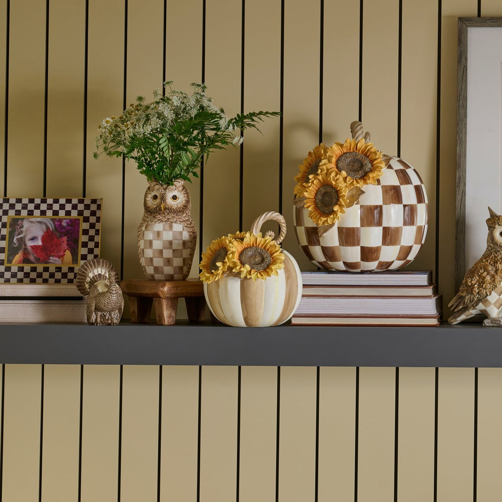 Decorative autumn-themed shelf display featuring checkered owl figurines, sunflower pumpkins, a small owl statue, and framed art against a beige paneled wall.
