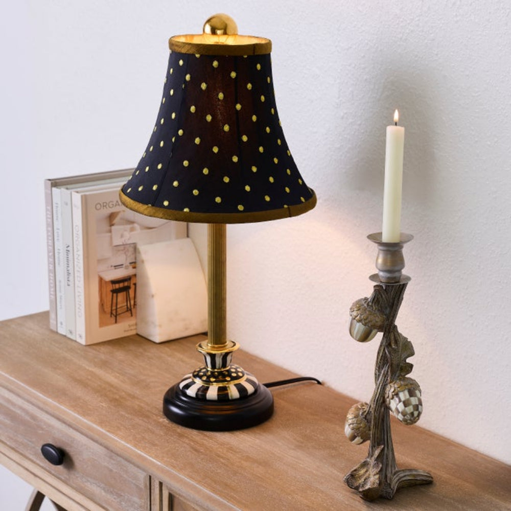 A table lamp with a black fabric shade covered in gold polka dots, a gold-toned stem, and a black-and-gold striped base, shown on a wooden table next to books and a candle holder with a lit candle.