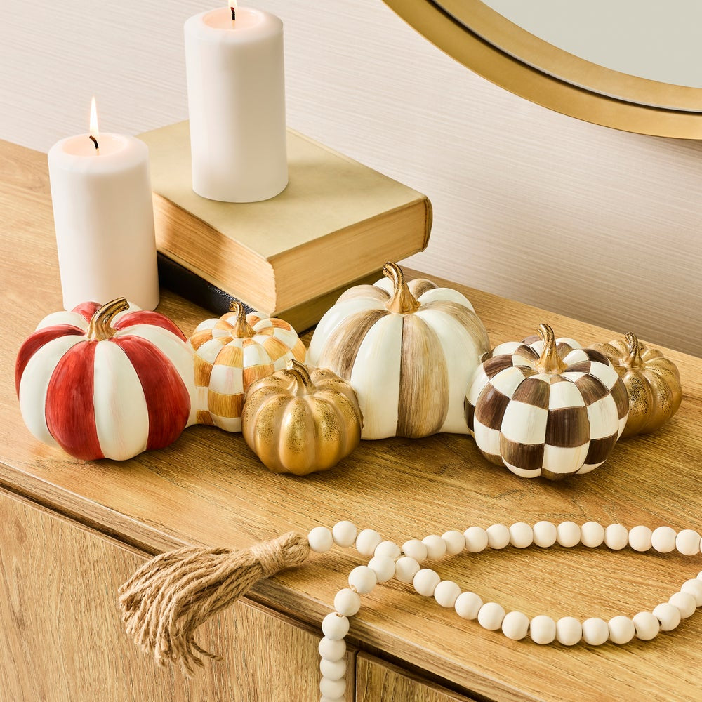 Decorative faux pumpkins in red-and-white, cream-and-cream, tan-and-cream, and checkered brown-and-cream patterns arranged on a wooden table beside white pillar candles and a book.