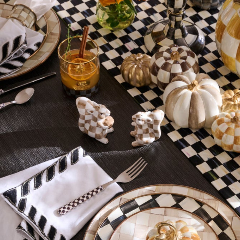 An autumn-themed table setting featuring a black and white checkered table runner, matching napkins, plates, and decorative pumpkins, with a glass of orange beverage on a dark wood table.