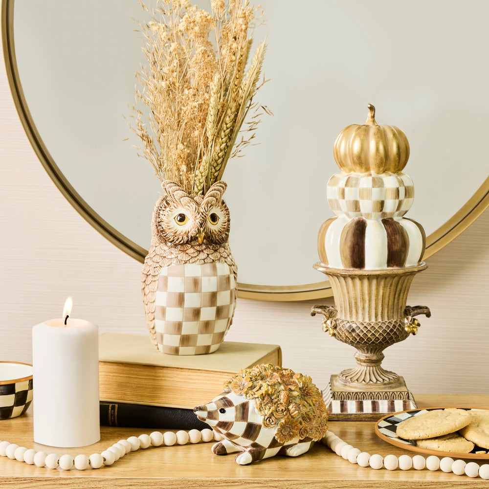 A styled tabletop scene with decorative ceramic owl and hedgehog figurines, a checkered candle holder, a candle, beads, cookies, a pumpkin-shaped vessel, and a round mirror in the background.