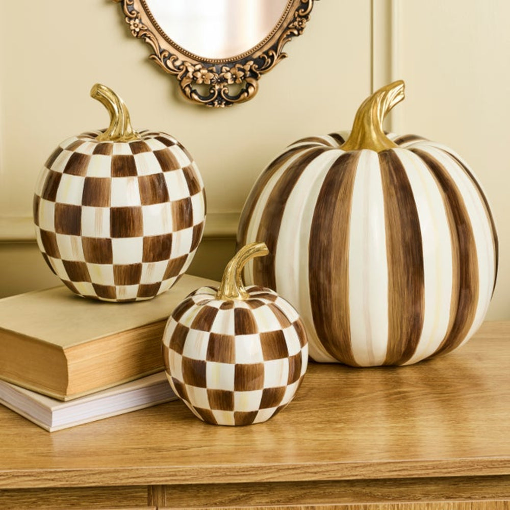 Three decorative pumpkins arranged on a wooden table with stacked books. Two pumpkins have a brown and cream checkerboard pattern, and one larger pumpkin has vertical brown and cream stripes.