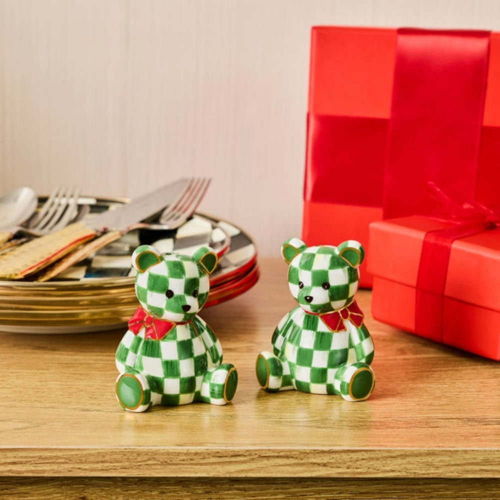 Two small teddy bear salt and pepper shakers with green and white checkered patterns and red bow ties, sitting on a wooden table next to wrapped red gift boxes.