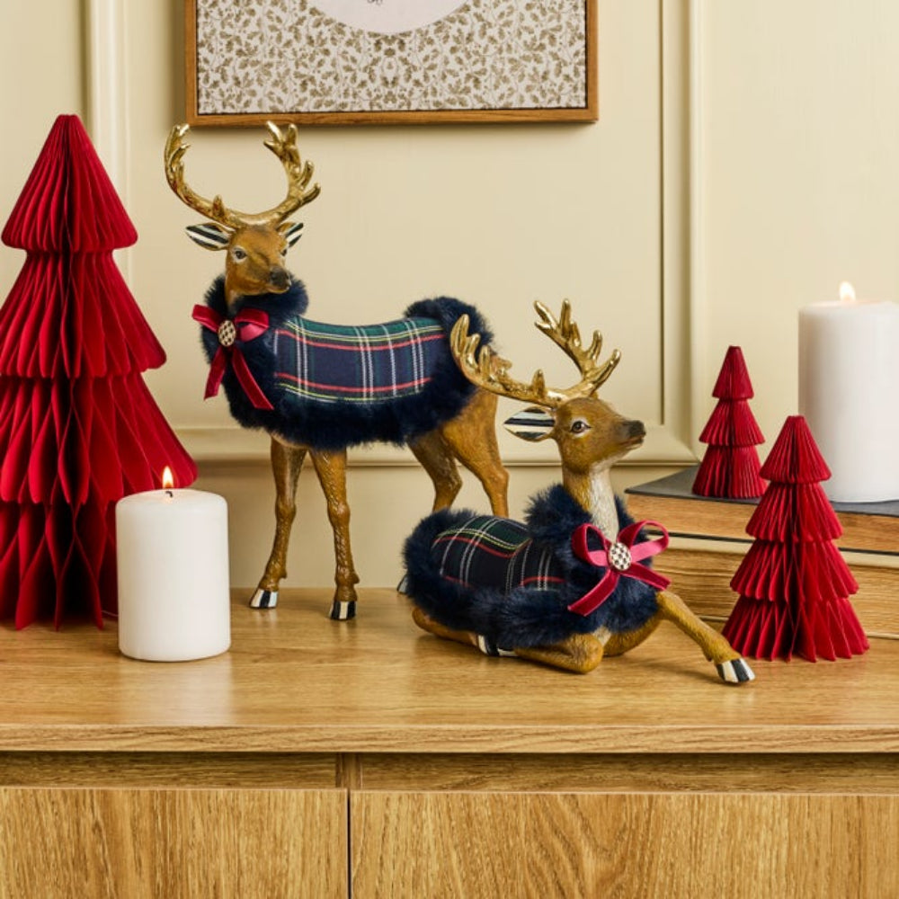 Two Christmas-themed deer figurines with gold antlers and navy blue faux fur, wearing tartan plaid scarves and red bows, displayed on a wooden cabinet alongside red paper trees and white candles.