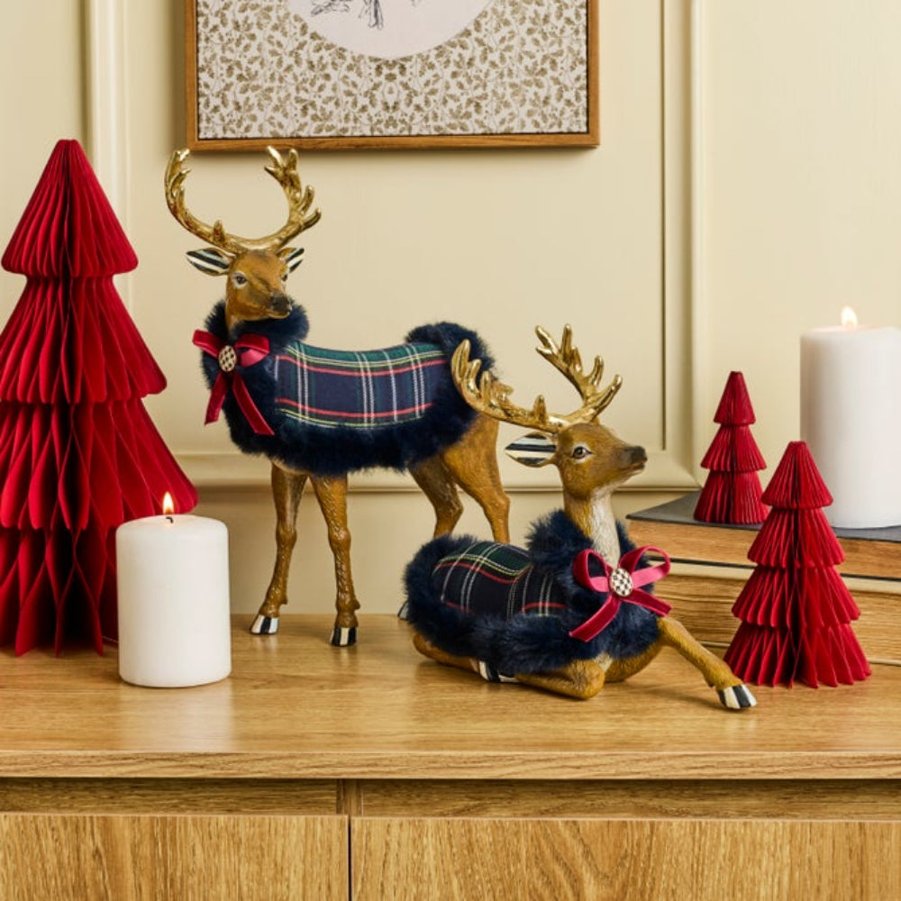 Two decorative deer figurines dressed in navy blue faux fur and tartan plaid scarves, styled on a wooden cabinet with red paper Christmas trees and white candles.