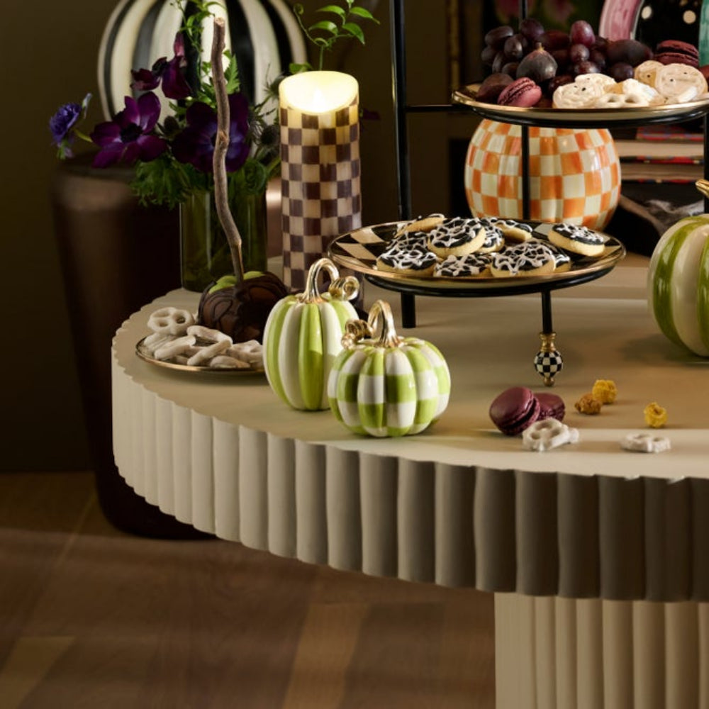 A styled tabletop scene with decorative striped pumpkins, a checkered candle holder, macarons, pretzels, and cookies on tiered stands in an indoor setting.