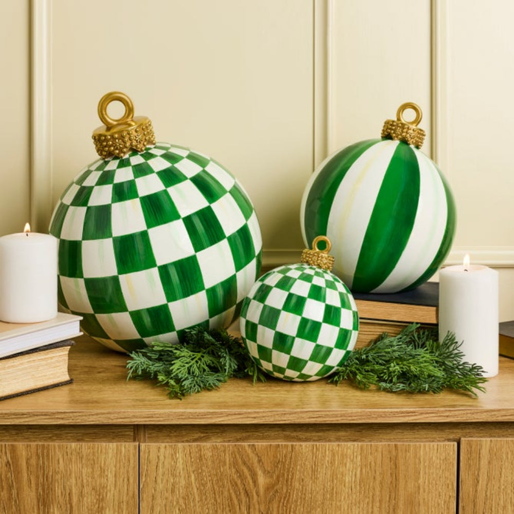 Three decorative Christmas ornaments in green and white patterns are arranged on a wooden cabinet with candles, books, and evergreen branches against a cream paneled wall.