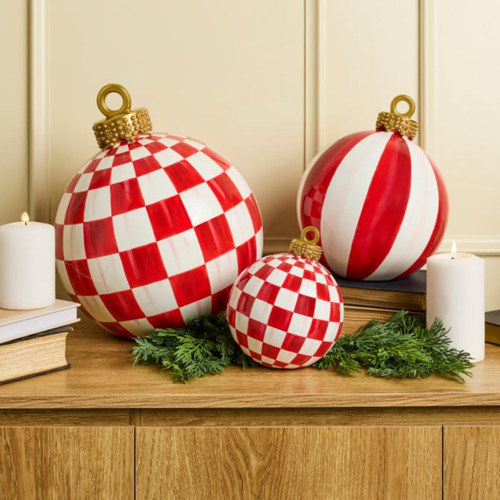 Three red and white checkered Christmas ornaments with gold caps, styled on a wooden cabinet with candles, books, and greenery against a beige paneled wall.