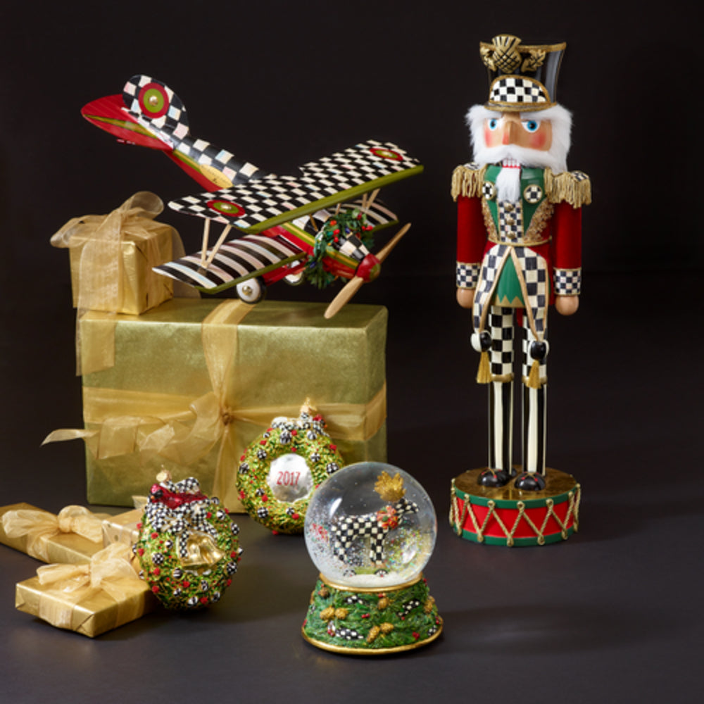 A festive holiday display featuring a tall nutcracker figurine, a red and black checkered toy airplane, a gold gift box with ribbon, a beaded ornament, a small snow globe, and wrapped presents on a dark studio background.