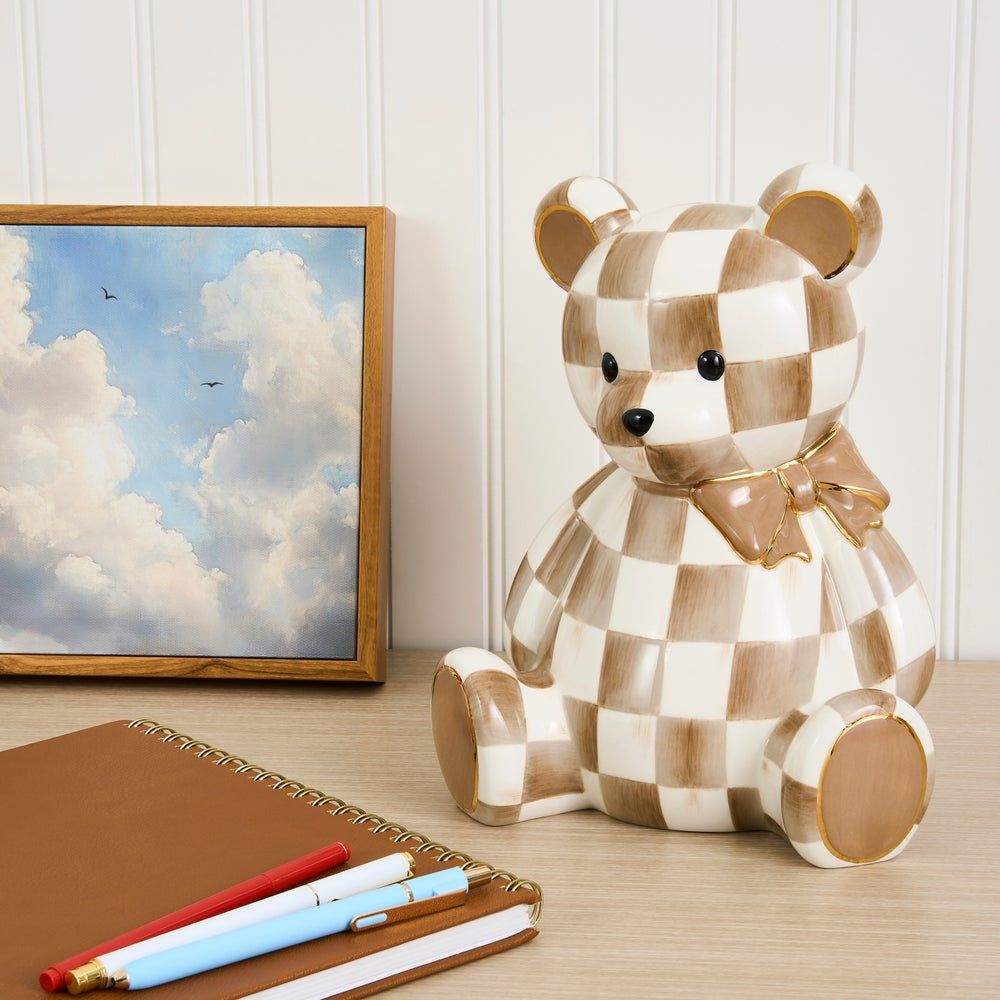 A ceramic teddy bear pig bank with a brown and cream checkered pattern and a matching bow tie, sitting on a wooden desk next to a framed picture, notebook, and pens.