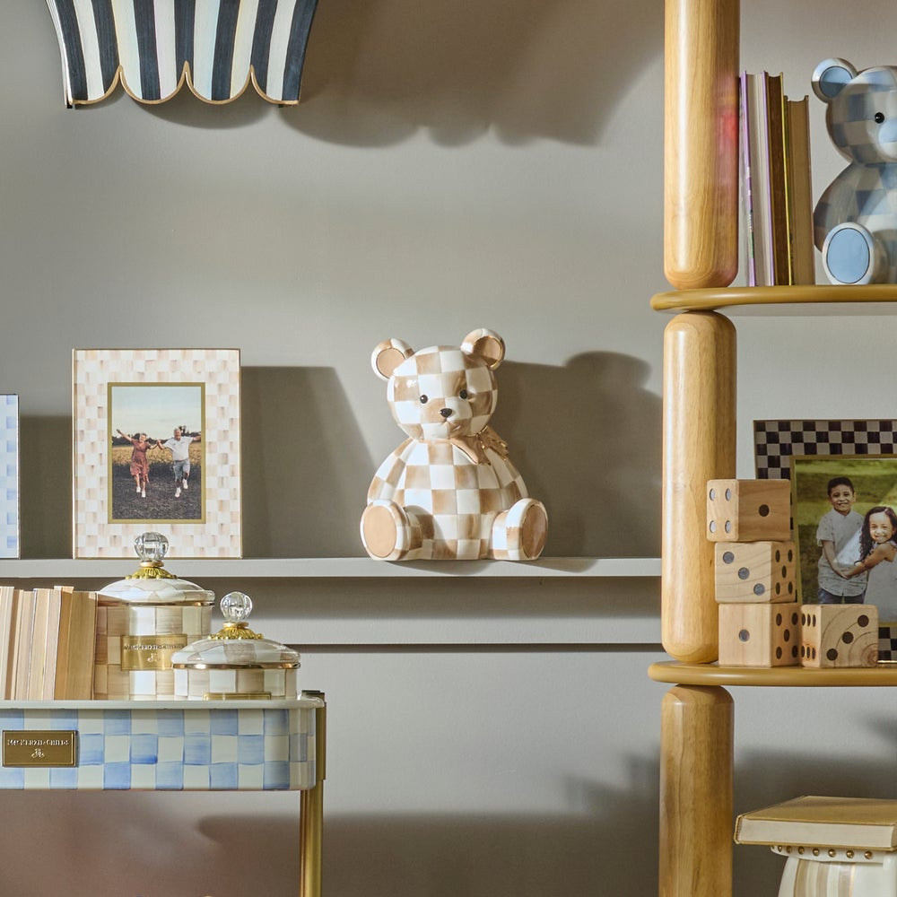 A beige and white checkered ceramic teddy bear figurine displayed on a shelf in a styled interior setting with wooden shelving, books, and picture frames.