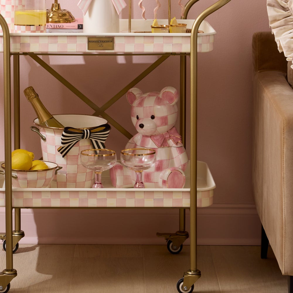Decorative bar cart with a teddy bear, glasses, and a bottle on a pink checkered tablecloth.