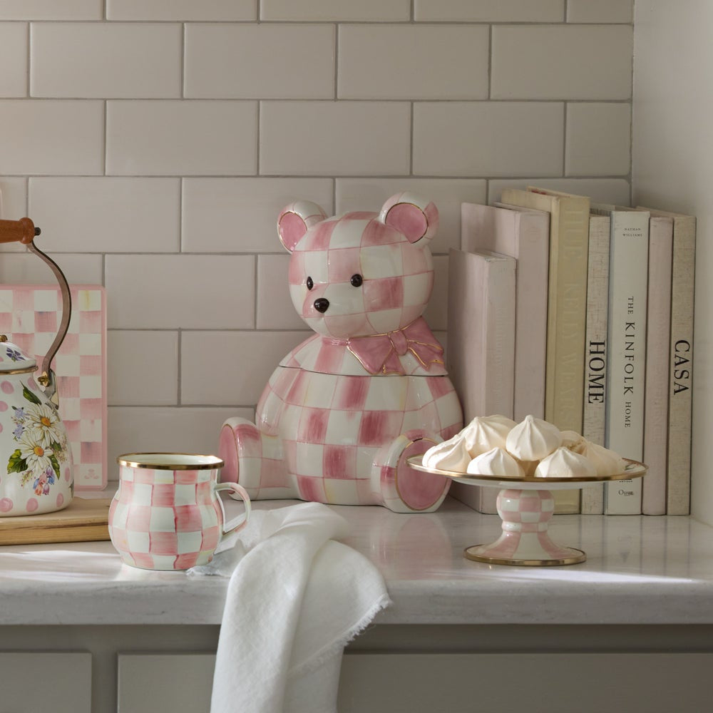 Checkered teddy bear, mug, and cake stand on a countertop with books in the background.