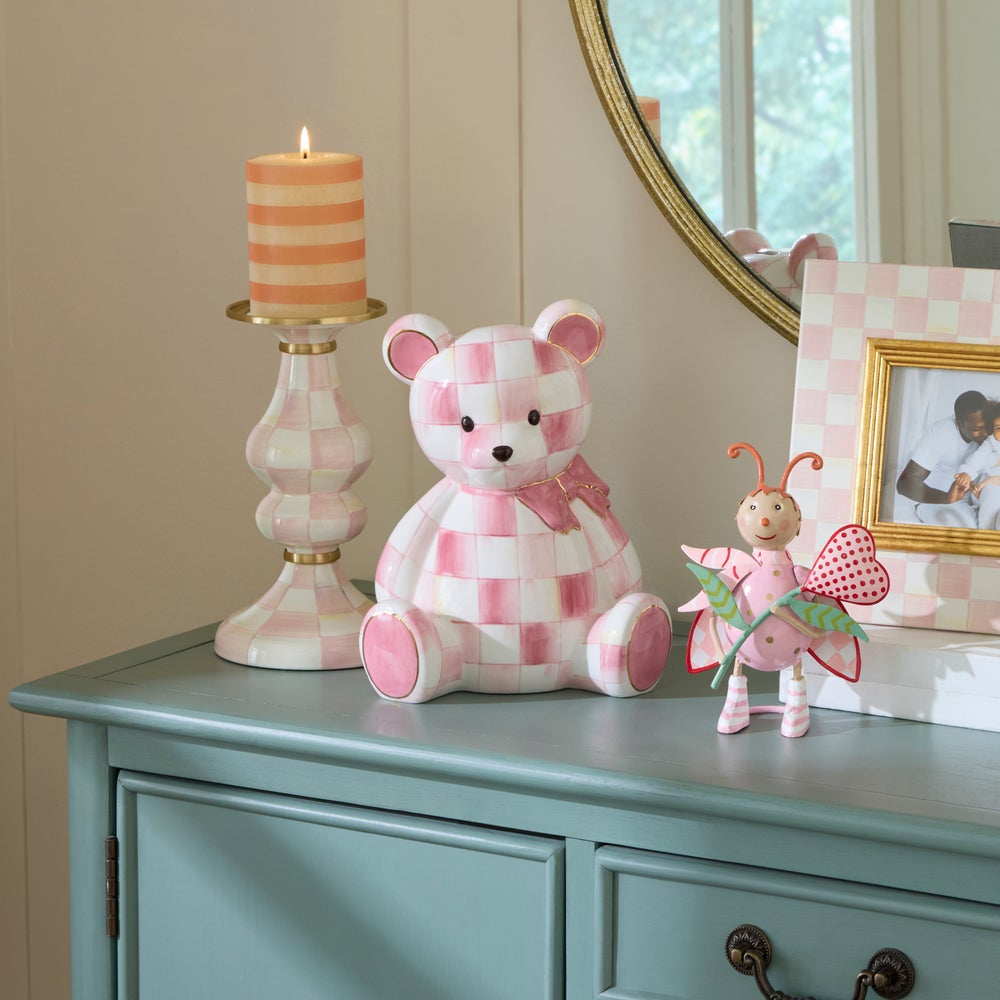 Checkered teddy bear and fairy figure on a dresser with a candle and mirror in the background.