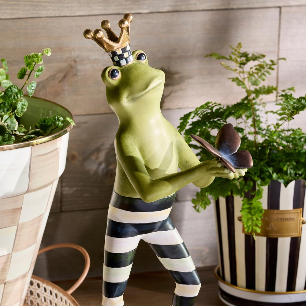 Decorative frog figurine with a crown holding a butterfly, standing between two potted plants.