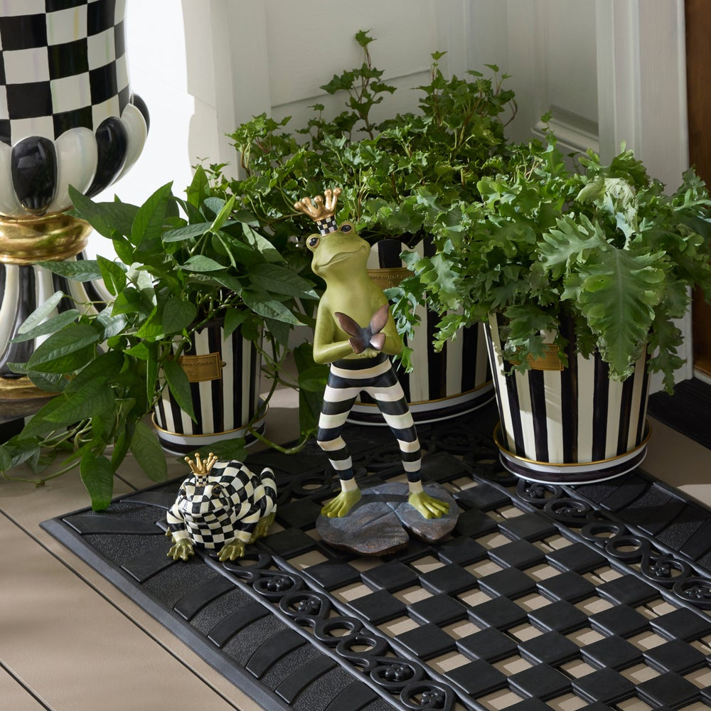 Decorative frog figurine with striped pants and a crown, surrounded by potted plants on a black doormat.