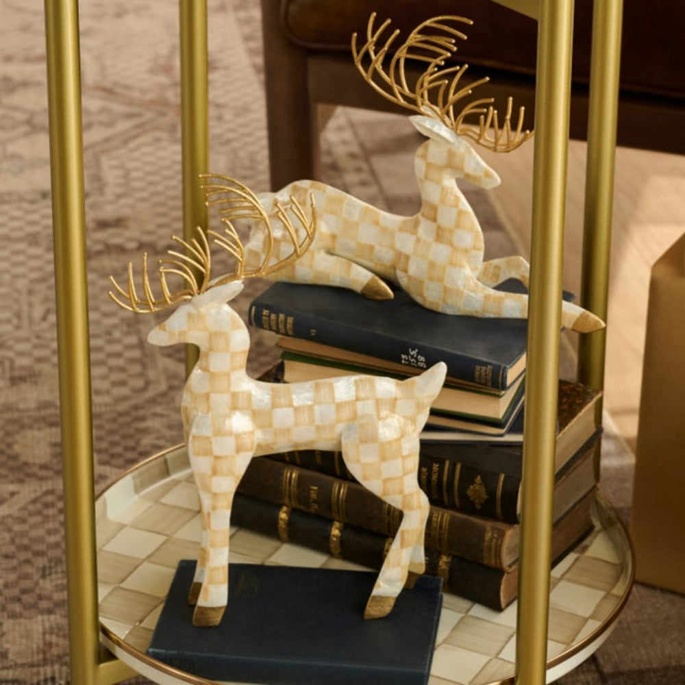 Two decorative deer figurines with a cream and gold checkered pattern and gold antlers, displayed on a round tray and stacked books beside a gold-framed side table.