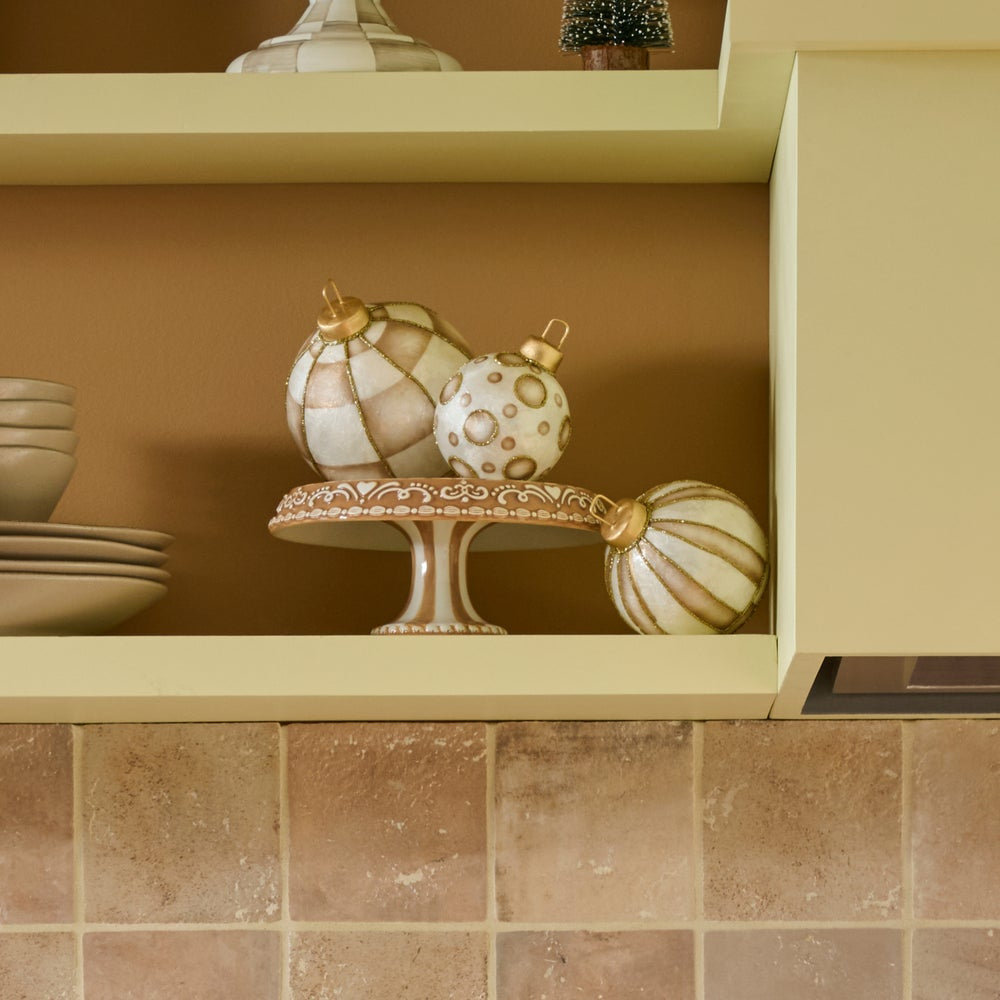 Three decorative Christmas ornaments in cream, beige, and white with gold accents displayed on a matching pedestal stand on a kitchen shelf.