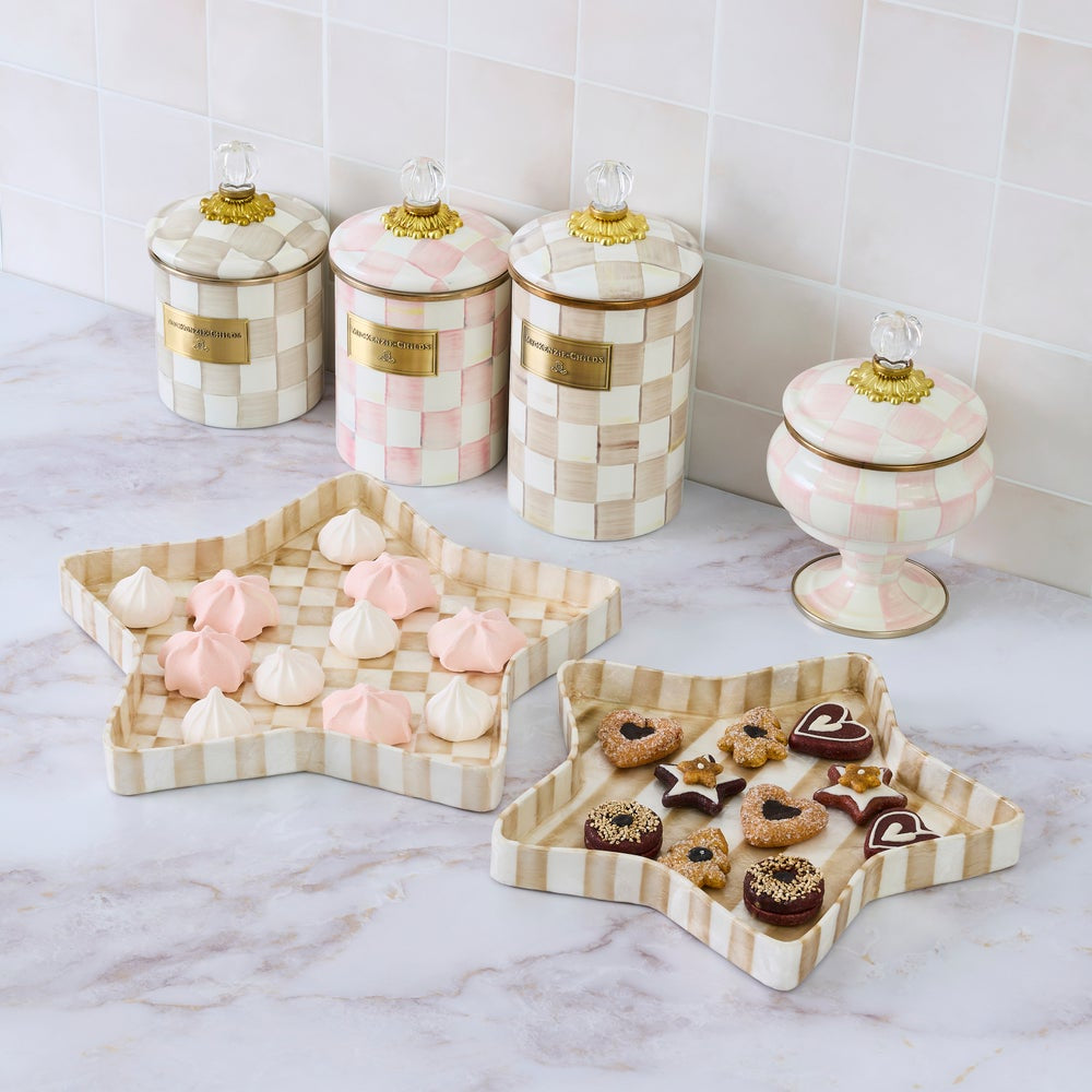 A pastel pink and cream checkered ceramic kitchen set displayed on a marble countertop against a tiled wall. The set includes three tall canisters, one pedestal lidded jar, a star-shaped tray, and a rectangular tray holding assorted meringues and chocolates.