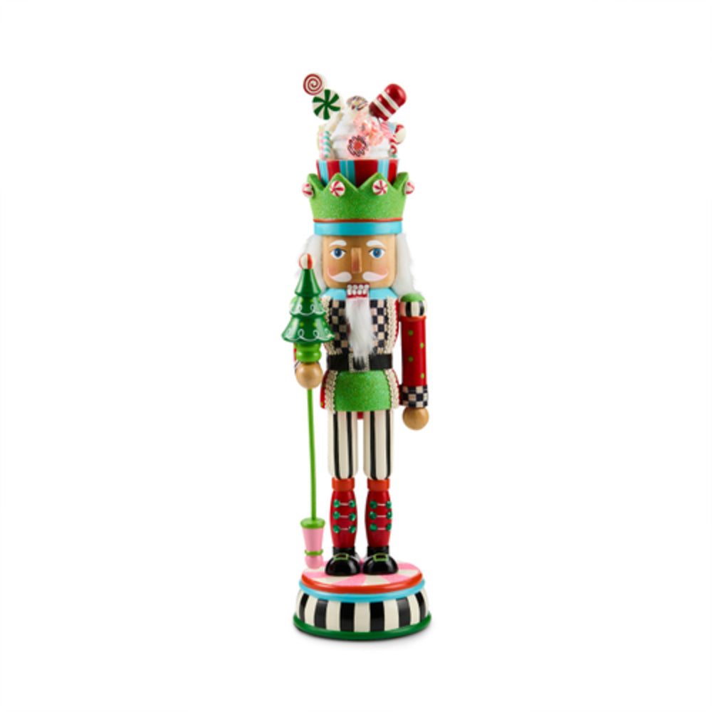 A festive Christmas nutcracker figurine with a green hat decorated with candy and a Santa hat, holding a small green Christmas tree, standing on a striped base against a white background.