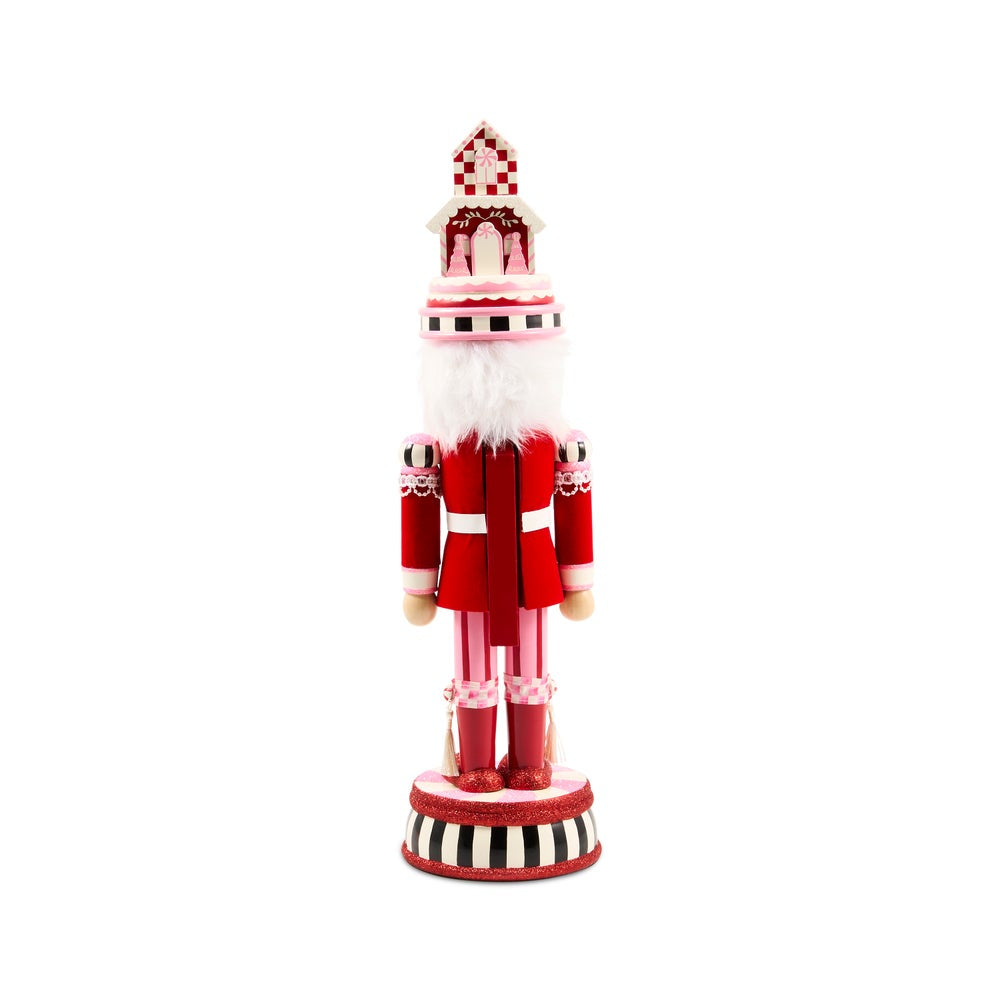 A festive red and white nutcracker soldier standing on a striped circular base, with a gingerbread house on its head, photographed against a solid white background.