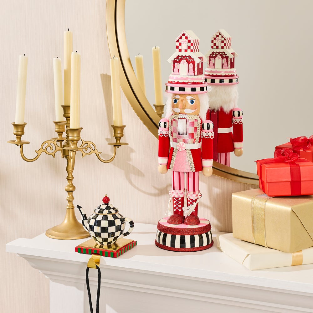 A red and white striped nutcracker figurine stands on a white mantel beside a gold candelabra, a checkered teapot, wrapped gift boxes, and a mirror reflecting its back.