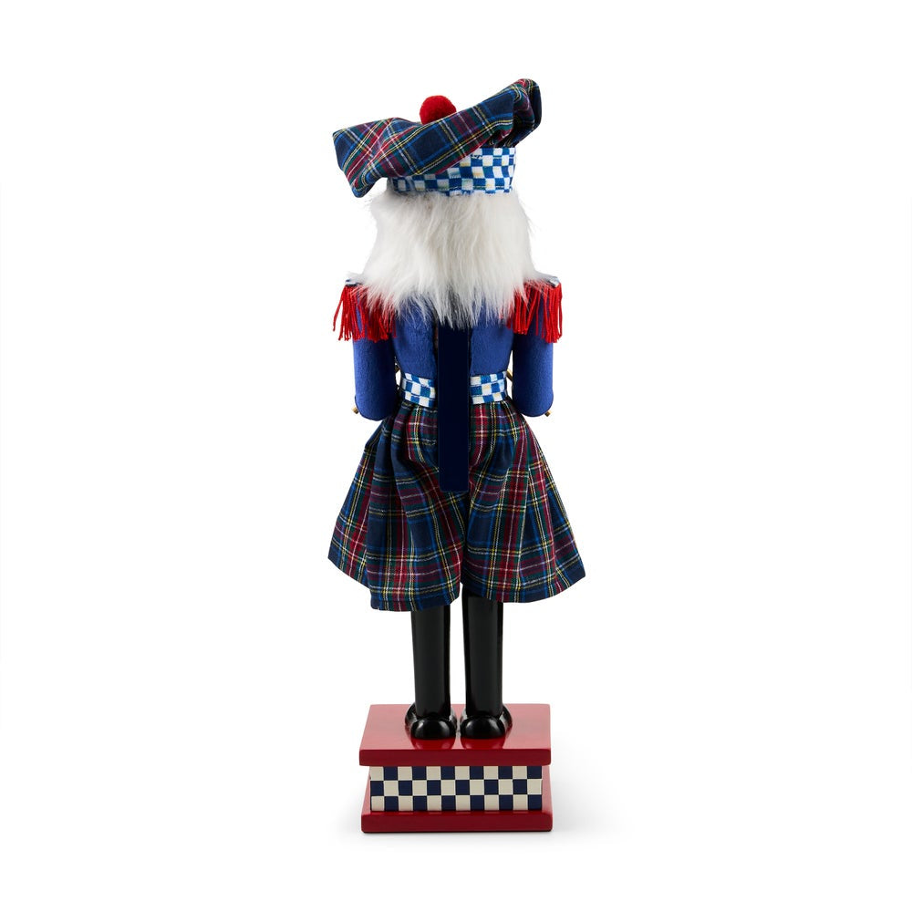Back of a Scottish kilted nutcracker figurine shown from the back, wearing a blue tartan kilt, matching hat, white faux beard, and red accents, standing on a red base against a white background.