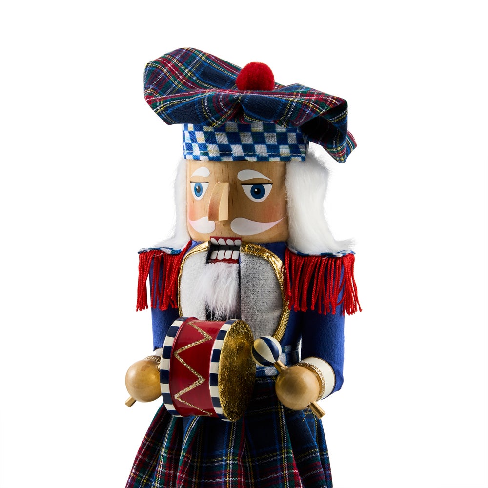 A traditional Scottish nutcracker doll with a blue tartan plaid hat and skirt, white hair, and a red pom-pom accent, holding a small drum against a white background.