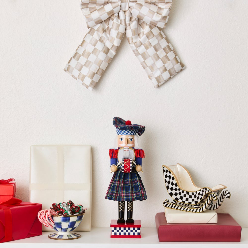 A festive holiday tabletop display featuring a central nutcracker figurine in a red, blue, and green tartan outfit, flanked by a beige-and-white checkered bow on the wall, wrapped gifts, candy, and a matching checkered shoe.
