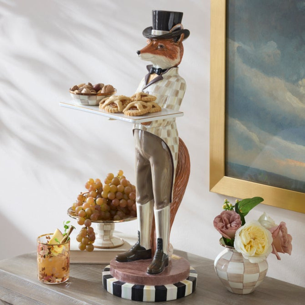 Decorative statue of a fox in formal attire holding a tray with food, placed on a surface with a drink and flowers.