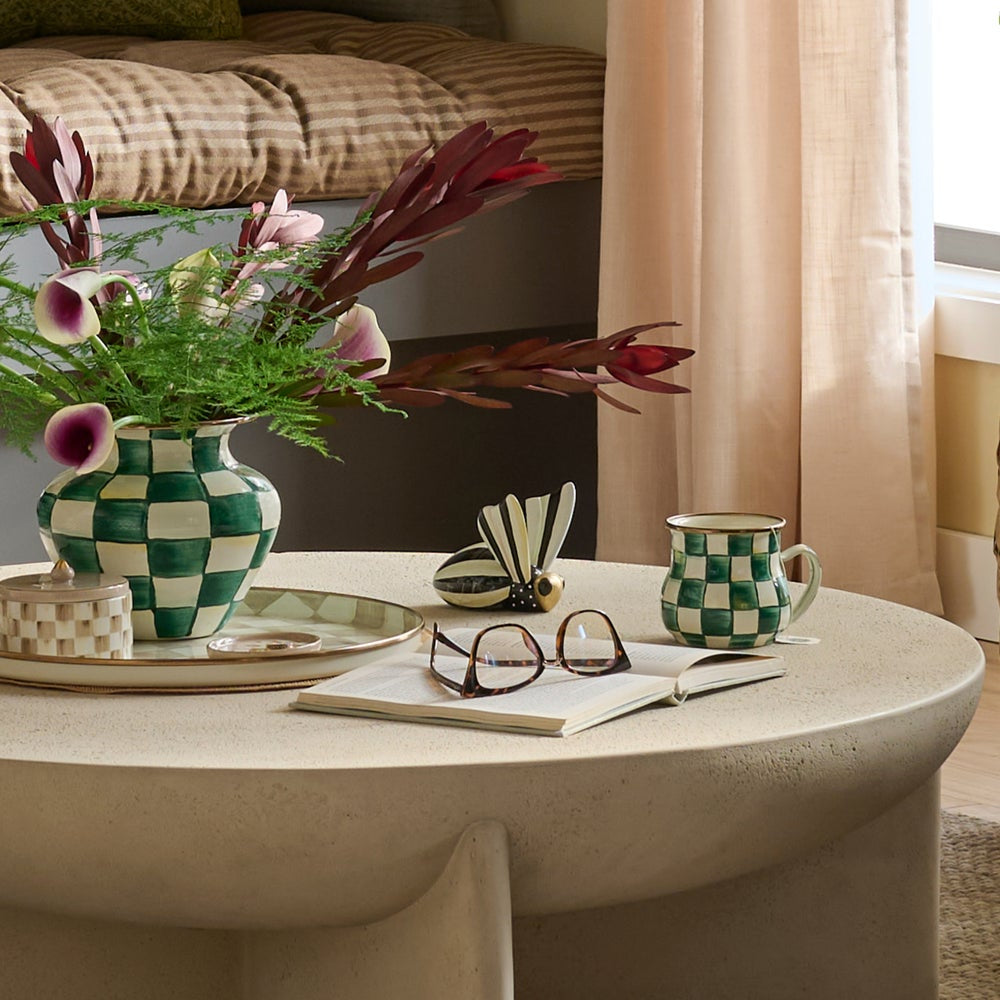A styled interior scene showing a round beige table with a green-and-white checkered ceramic vase of flowers, a matching checkered mug, eyeglasses, and an open notebook in a softly lit room.