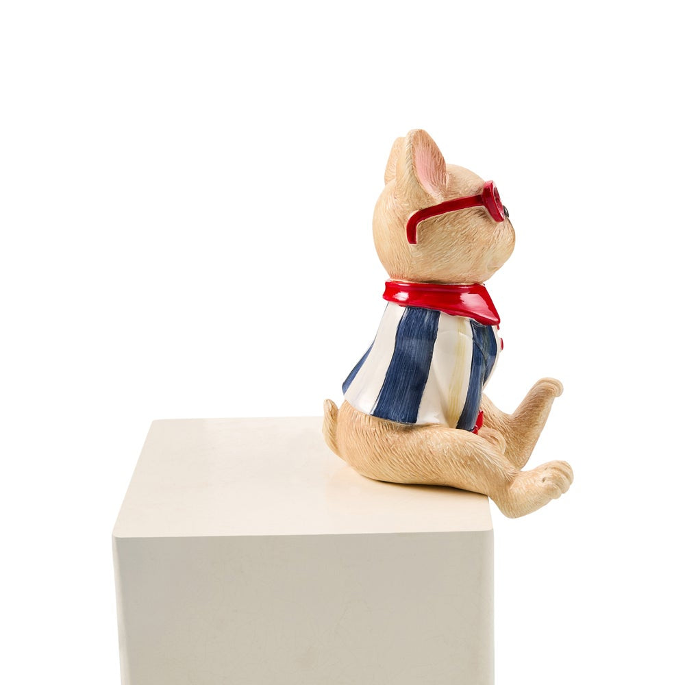 A small beige plush toy or figurine of a cat sitting on a white block, wearing a blue and white striped shirt, a red neck scarf, and red sunglasses, photographed against a white background.