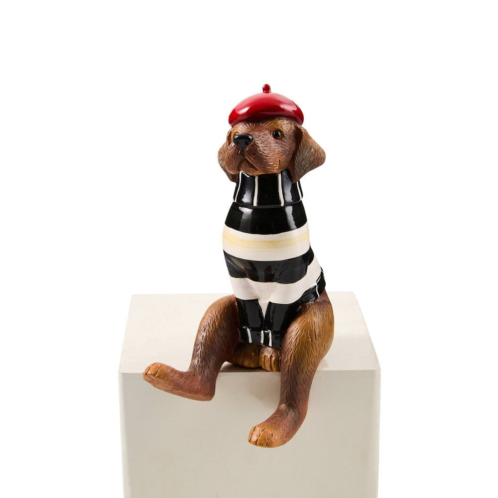 A dachshund figurine wearing a red beret, a black and white striped sweater, and black pants, sitting on a white block against a white background.