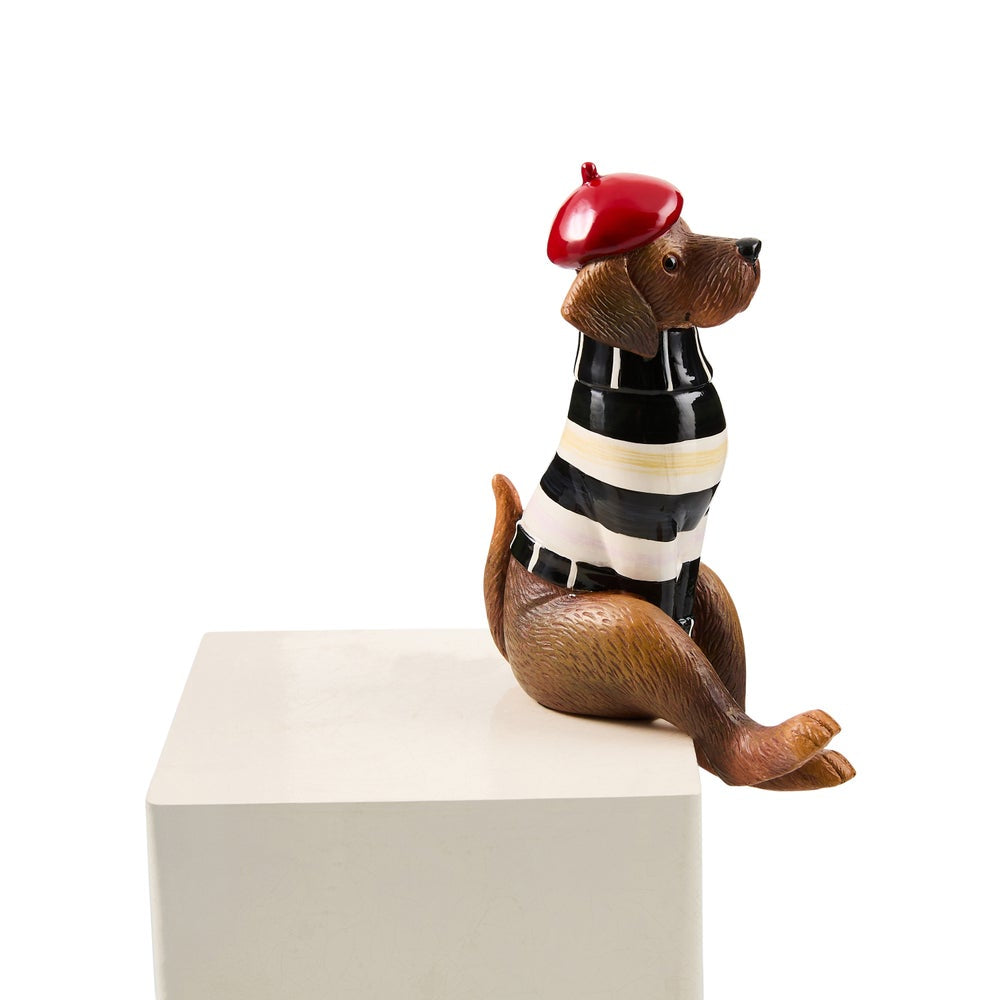 A decorative figurine of a seated brown dog wearing a red beret, a black-and-white striped shirt, and a black-and-white striped belt, displayed on a white pedestal against a white background.