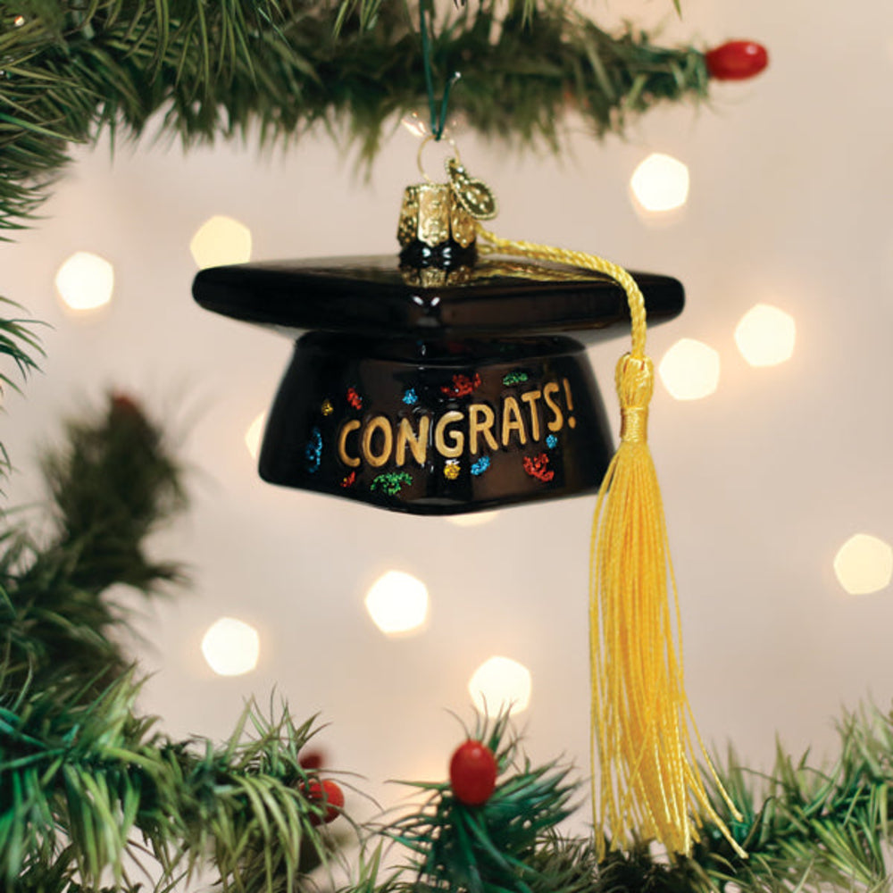 A black graduation cap Christmas ornament hanging from a Christmas tree branch, with 'CONGRATS!' in gold lettering and a yellow tassel, against a warm bokeh background.