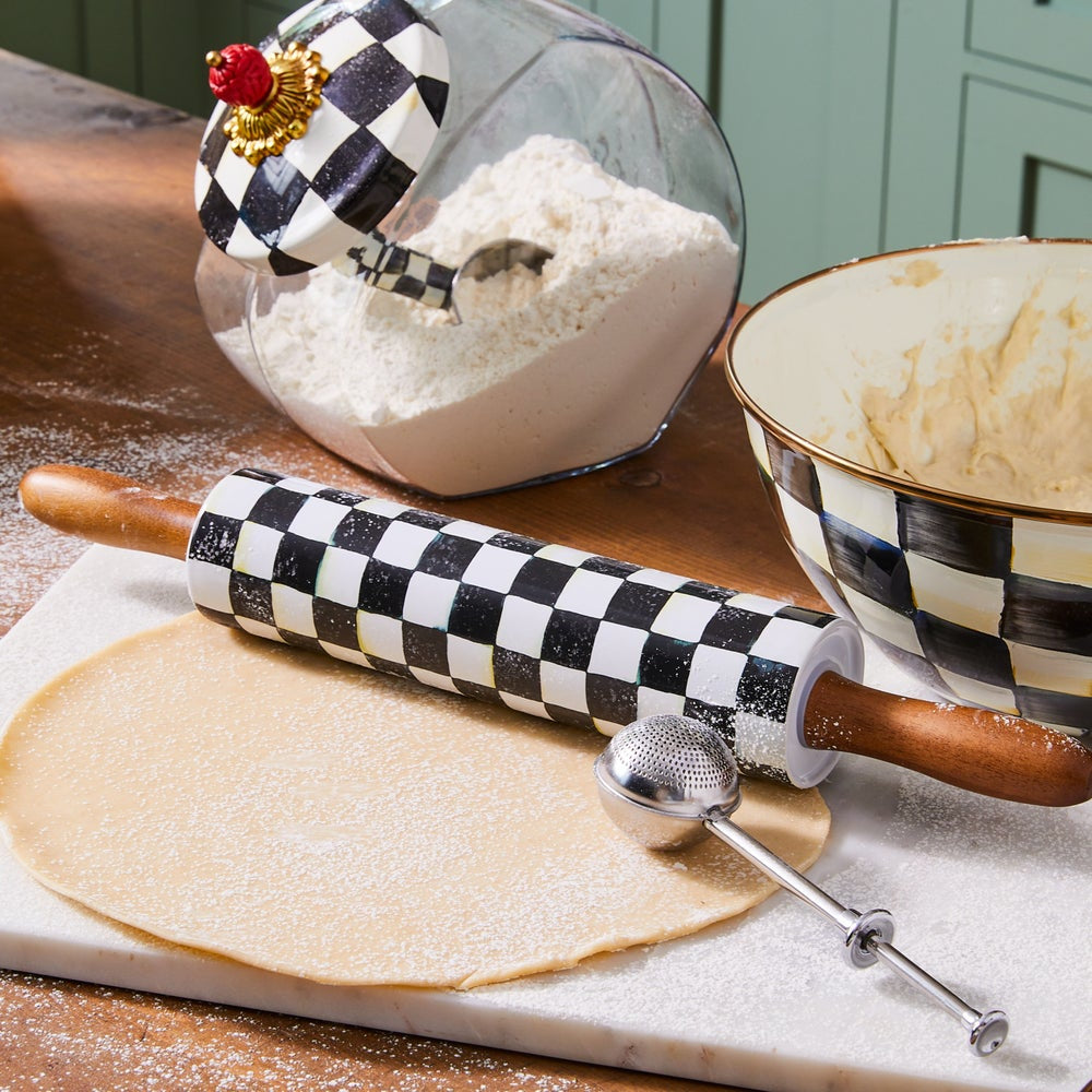 Baking setup with checkered rolling pin, bowl of flour, and rolled-out dough on a wooden surface.