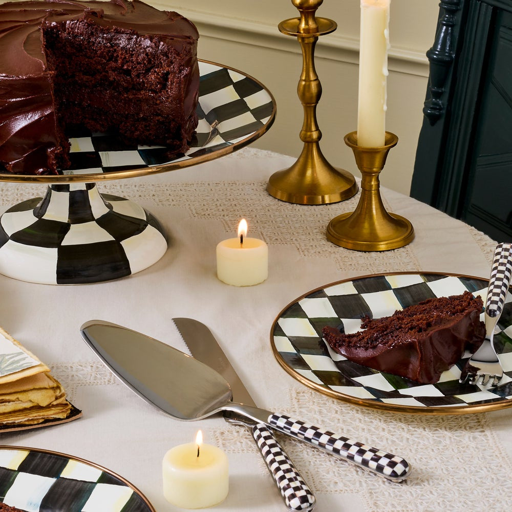 A styled tabletop scene with a slice of chocolate cake on a black-and-white checkered plate, a whole cake on a matching pedestal plate, cake servers with black-and-white checkered handles, candles, and gold candlesticks on a white tablecloth.