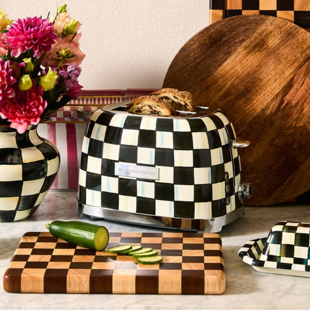Checkered toaster with bread, cutting board with cucumber, and flowers on a checkered tablecloth.