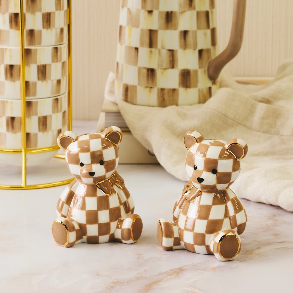 Two ceramic teddy bear figurines with a brown and cream checkered pattern, sitting on a light marble surface next to a gold wire stand and a beige towel.