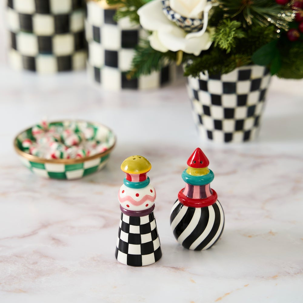 Two decorative salt and pepper shakers with black-and-white checkered and striped patterns, topped with colorful candy-like accents, sit on a light marble surface. A matching checkered bowl of candy and blurred checkered vases with flowers appear in the background.