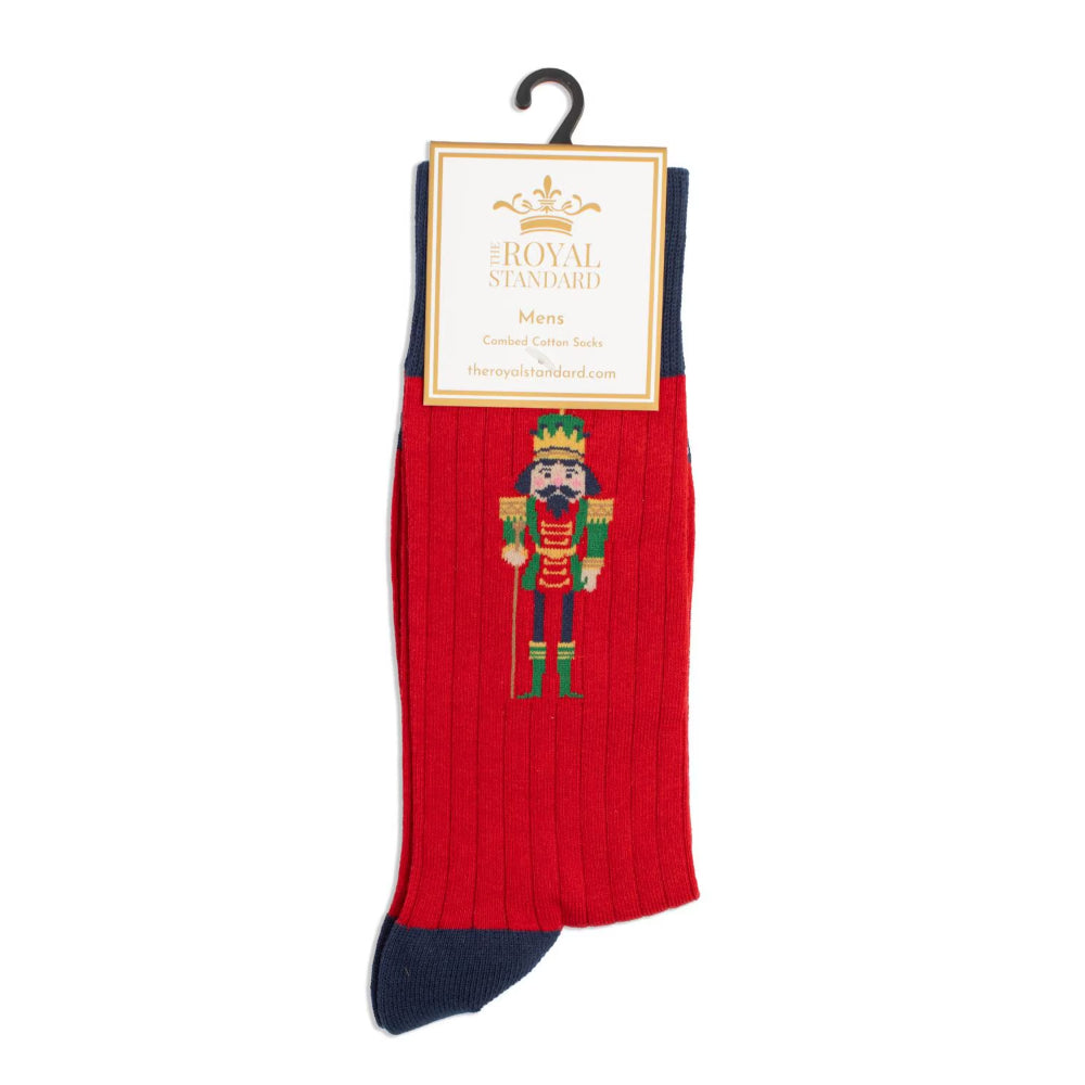 A single red ribbed crew sock with a navy blue toe and heel, featuring an embroidered nutcracker design on the front. The sock is displayed on a black hanger with a cream paper label that reads 'The Royal Standard'.