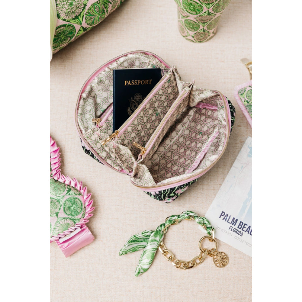 A patterned fabric zippered pouch with a beige and green monogram-style print and pink trim, shown open with a passport inside. The pouch is styled on a light beige surface with matching accessories including a scarf, bracelet, and other patterned items around it.