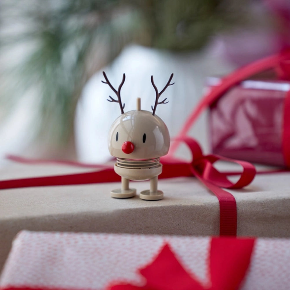 A small beige reindeer figurine with dark brown antlers and a red nose stands on a gift box wrapped in beige paper with a red ribbon, with blurred Christmas decorations in the background.