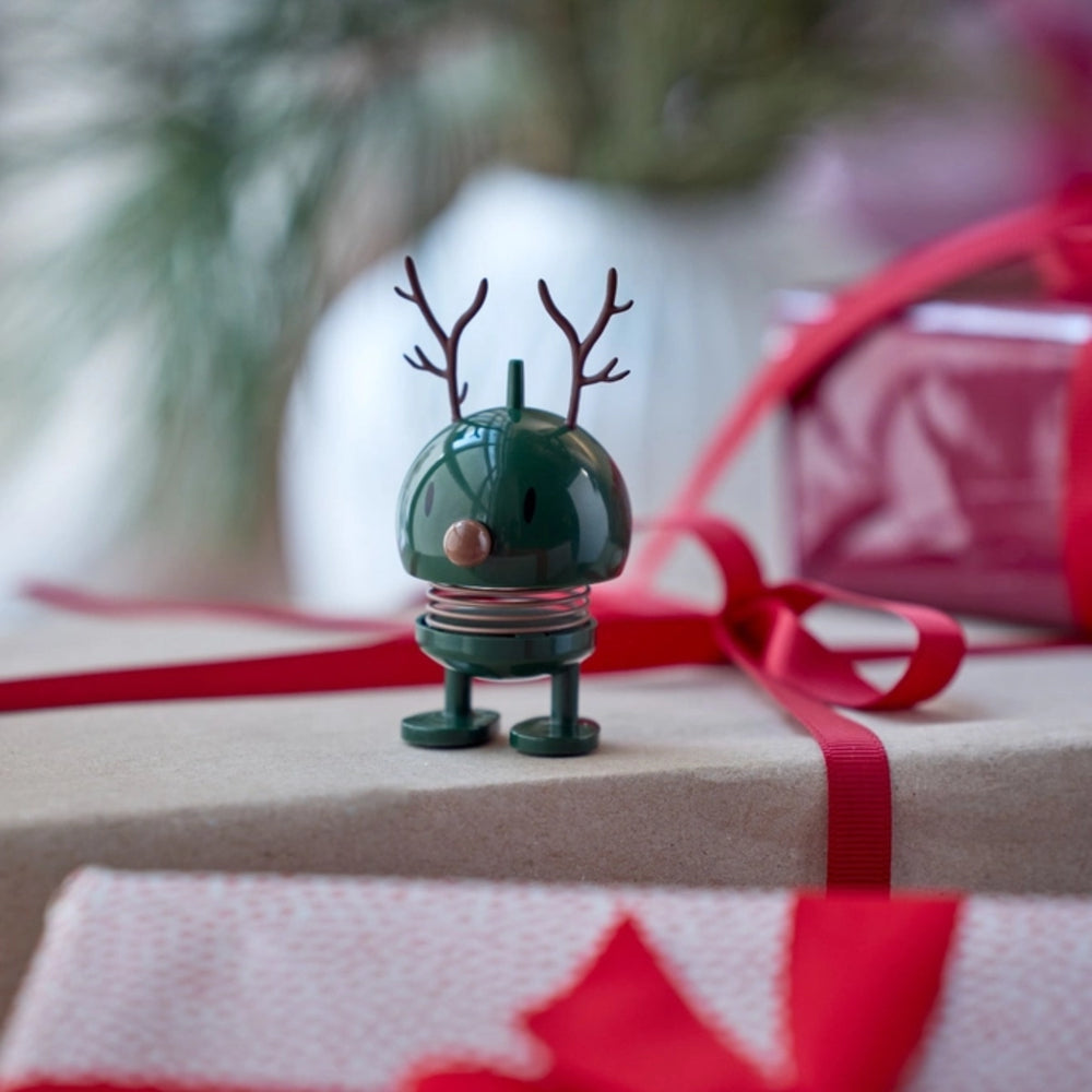 Small green reindeer figurine on a wrapped gift with red ribbon and blurred Christmas tree in the background.