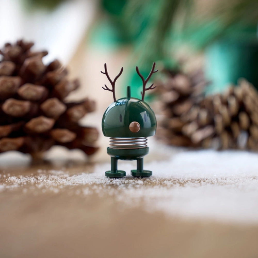 Small green figurine with antlers on a snowy surface with pinecones in the background.