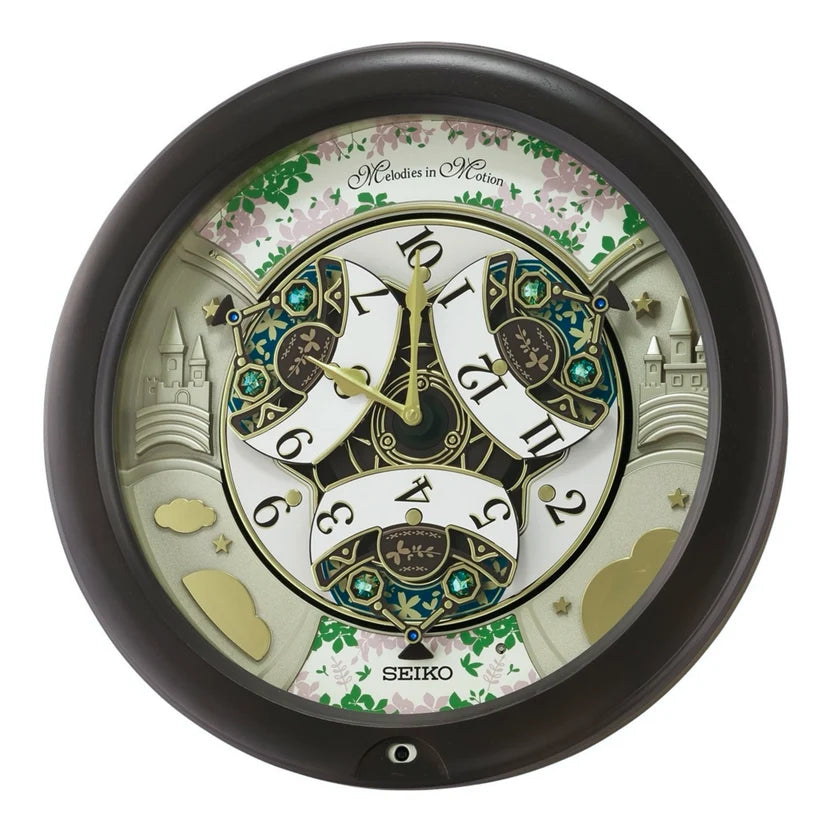 A Seiko wall clock with a black frame and a highly detailed, whimsical clock-face dial. The dial features a fantasy garden theme with illustrations of castles, flowers, and green leaves, alongside stylized clock hands and Roman numerals on white banners.