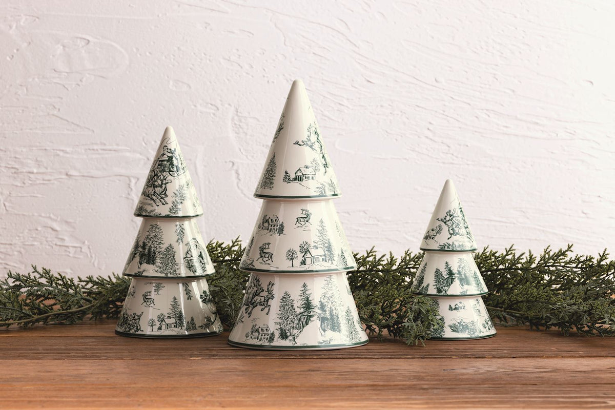 Three tiered Christmas trees with a green toile de Jouy winter landscape pattern on an off-white background, arranged on a wooden surface with green pine garland against a white textured wall.