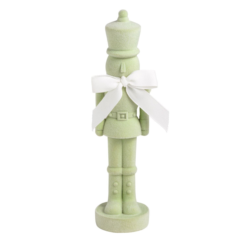 A light sage green nutcracker figurine with a white ribbon bow around its neck, standing on a round base against a white background.